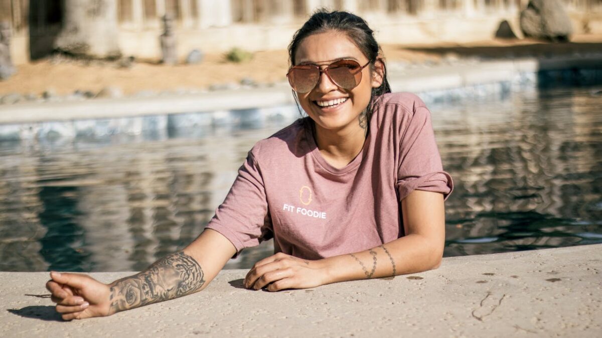 Smiling woman with sunglasses and tattoos enjoying a sunny day by the pool.